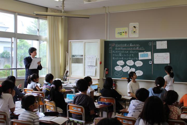 授業をする先生と子どもたちの横で見学している教育委員の写真