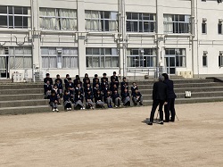 校舎の前の階段に並んで座っている制服姿の生徒たちと、三脚でカメラを構える2人の男性の写真