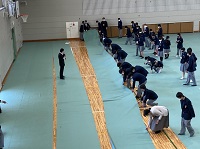 体育館の床に緑の大きな布を敷いている、制服姿の生徒たちの写真