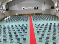 整然と並べられた数多くのパイプ椅子の中央に赤いレッドカーペットが敷かれた体育館を、後方から映した写真