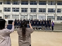校舎の前で制服姿で横数列に並ぶ生徒たちを2名の女性がタブレットで撮影している様子の写真