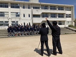 校舎の前で制服姿で横数列に並ぶ生徒たちに向かい三脚でカメラを構えた男性とその脇で手をあげ合図する男性の写真