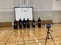 体育館で剣道着姿の6人が三脚に設置されたタブレットに向かい立っている写真