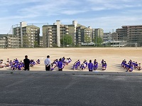 グラウンドに紫色のジャージを着た生徒たちが並んで座っている様子の写真
