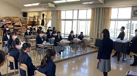 段差のある教室で生徒たちが並んで座り、その前に一人の女子生徒が立っている様子の写真