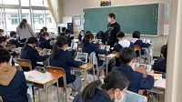 教室に生徒たちが並んで机に座り、黒板の前に男性が立っている様子の写真