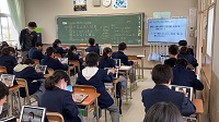 黒板に文字がたくさん書かれていて、その右側のモニターに資料が表示されていて、生徒たちが机に座っている教室の写真