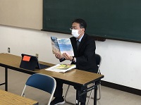 スーツ姿の男性が、黒板の前の長机に座り、青い冊子を見ながら話している様子の写真