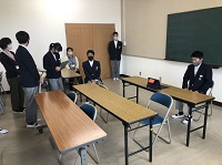 黒板の前の長机に制服を着た男子生徒が座り、向かいに他の生徒や先生がいる様子の写真