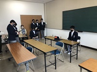 黒いマスクをした制服を着た生徒が、長机に座り、その右側の奥に生徒たちと先生が見守っている様子の写真