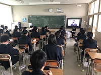 黒板に沢山の文字が書かれている教室で生徒たちが机に座り、左側に設置されたモニターを見ている様子の写真