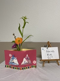 ひな祭りをモチーフにした紙のプランターにオレンジ色の花と紫色の花が特徴的な生花の写真