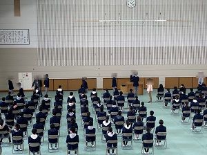 薄水色のシートが敷かれた体育館に並べられたパイプ椅子に座っている生徒たちの写真