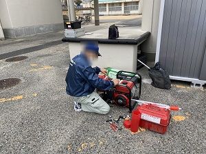 濃紺のジャンパーを着た男性がしゃがみ込み、赤い発電機を点検している様子の写真