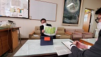 室内でテーブルを挟んで座り、緑色に塗られた紙を見せたりテーブルのメモを見ている2人の男性の写真