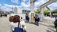 校門前の入学式と書かれた立て看板の横でピースサインをしている生徒たちと、それを携帯で撮っている2人の女性の写真
