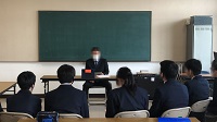 黒板の前に一人の男性が座りその前に向かい合うように数人の生徒が扇形に並べたパイプ椅子に座っている様子の写真