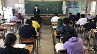 教室に生徒達が机に向かって座り、黒板の前にスーツ姿の男性が立っている様子の写真