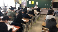 生徒たちが教室で机に向かって座り、黒板の前にマスクをした白いシャツを着た男性が座っている様子の写真