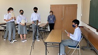 三脚に固定されたカメラの前に設置された机に座り原稿を読み上げている様子の生徒の写真