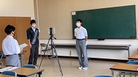 三脚に設置されたカメラの前にマスクをした男子生徒が立っている様子の写真