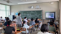 生徒のみんなが座っている教室で、灰色の服を着た生徒の一人が立っている様子の写真