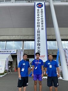 陸上競技大会の垂れ幕の前で青い服を着た選手と監督の写真