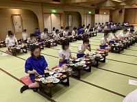 畳張りの宴会場で白と青のジャージ服姿の生徒たちが前後にきれいに並んで座り食事をしている様子の写真