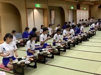 畳張りの宴会場で白と青のジャージ服姿の生徒たちが一方向を向いてきれいに並んで座り食事をしている様子の写真