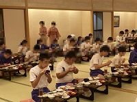 食事をする生徒たちの後ろでホテルの中居さん達が仕事をしている様子の写真
