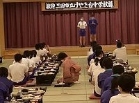 「歓迎三田市けやき台中学校様」と大きく書かれた横断幕のかかる舞台で2人の生徒たちが挨拶をしている様子の写真